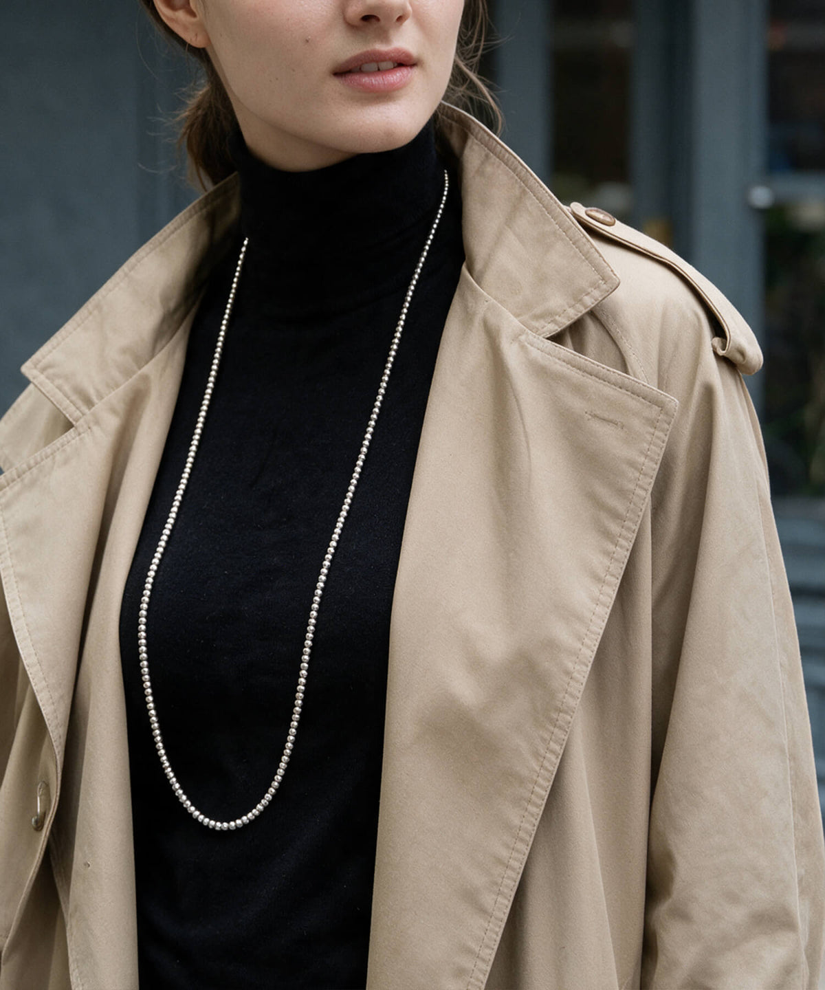 Long silver ball chain necklace worn doubled over a black turtleneck and trench coat