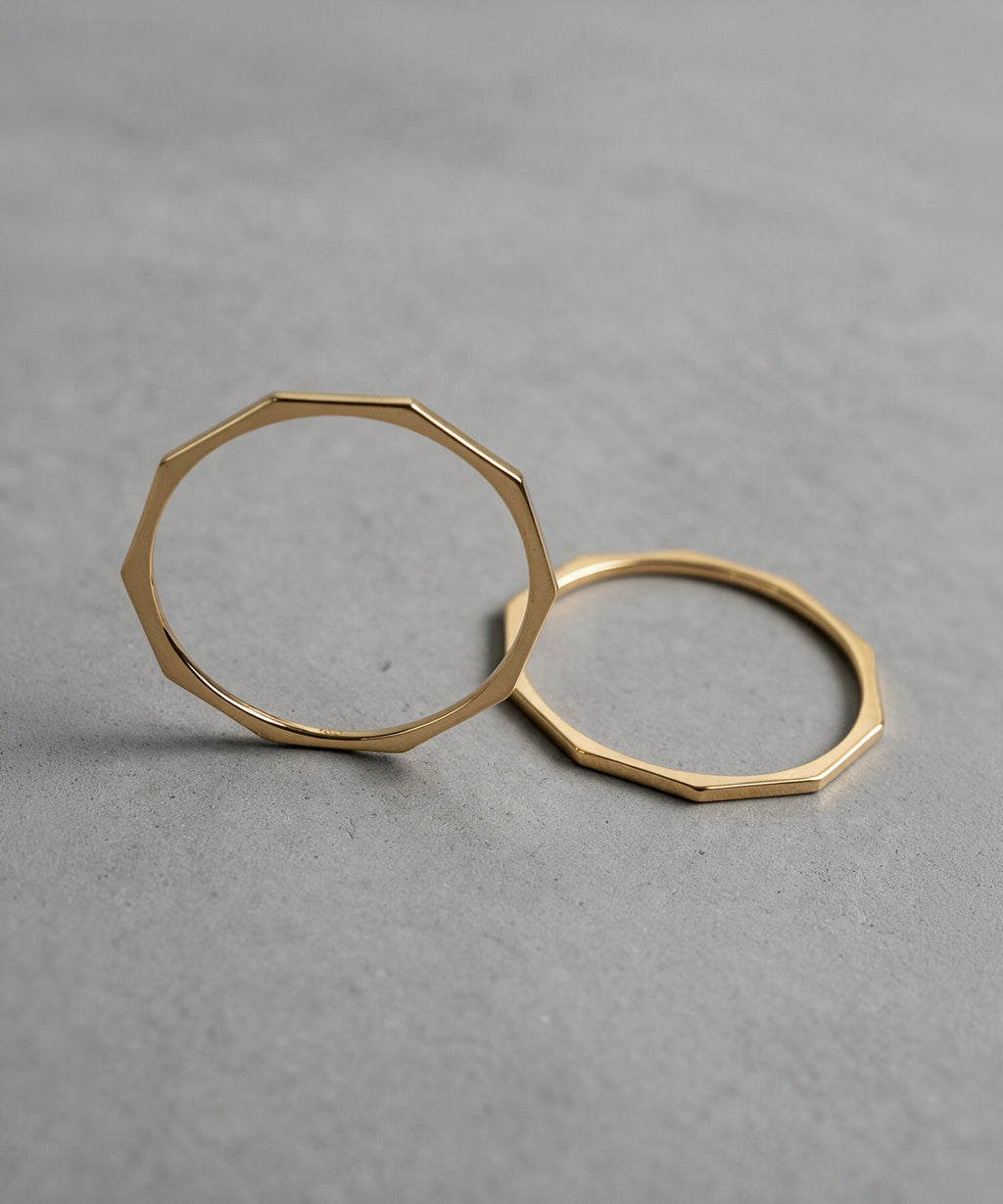 Two 18K gold decagon skinny rings laid on a grey surface