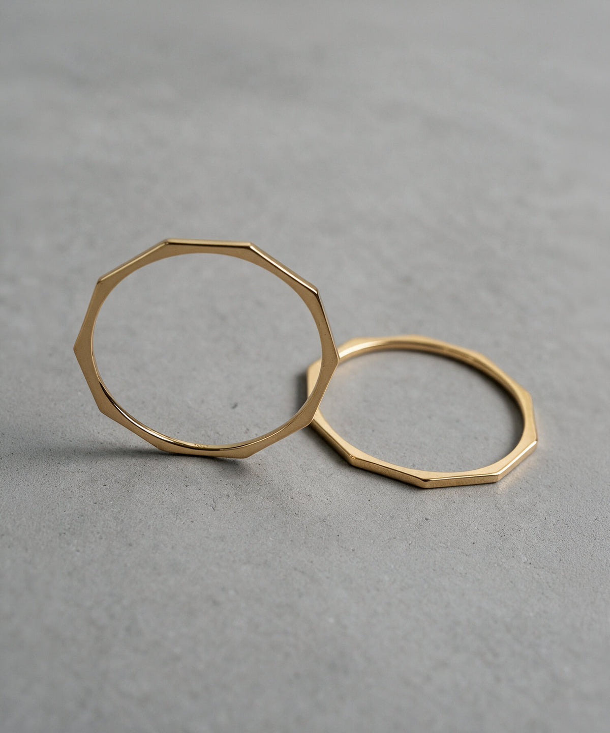 Two 18K gold decagon skinny rings laid on a grey surface